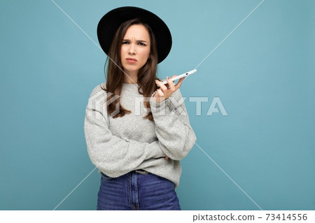 Beautiful young brunette woman thinking wearing black hat and grey sweater holding smartphone 73414556