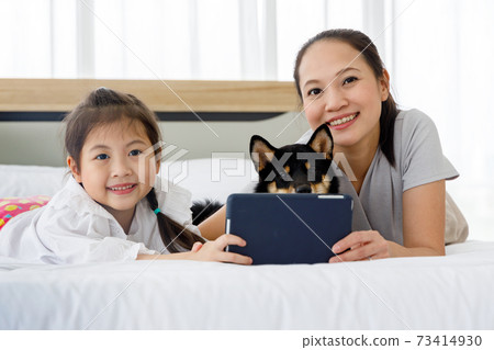 Happy Asian mother and daughter lying on bed and using digital tablet. Black Shiba Inu on bed between its owner. Concept family with dog at home. Happy Asian mother and daughter lying on bed and using digital tablet. Black Shiba Inu on bed between its owner. Concept family with dog at home. 73414930