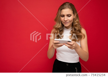 Photo of concentrated beautiful young blonde woman wearing white t-shirt isolated on red background 73415234