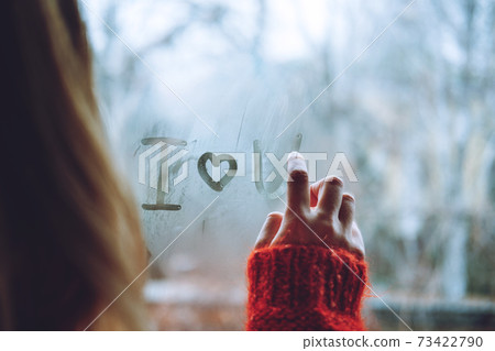 Valentines Day During The Coronavirus Outbreak. Love at distance, loneliness in self-isolation in the time of coronavirus. Woman draws heart and I love you standing by the window at home Valentines Day During The Coronavirus Outbreak. Love at distance, loneliness in self-isolation in the time of coronavirus. Woman draws heart and I love you standing by the window at home 73422790