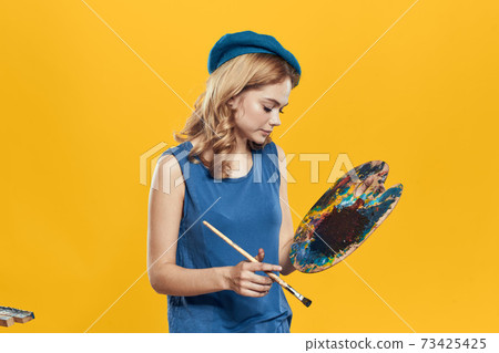 Woman with palette in blue hands takes paintbrush hobby artwork yellow background 73425425
