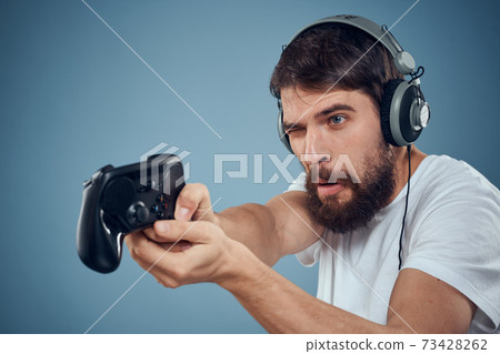 Man in headphones controller in hands playing game console lifestyle emotions blue background white t-shirt Man in headphones controller in hands playing game console lifestyle emotions blue background white t-shirt 73428262