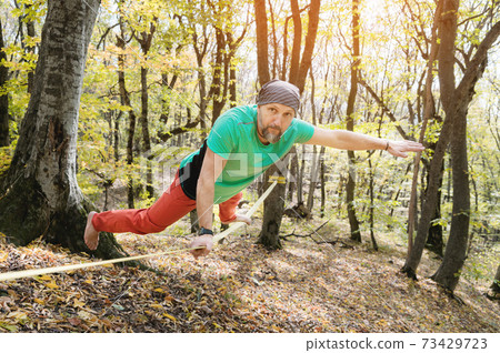 A bearded man in age balances while sitting on a taut slackline in the autumn forest. Outdoor Leisure A bearded man in age balances while sitting on a taut slackline in the autumn forest. Outdoor Leisure 73429723