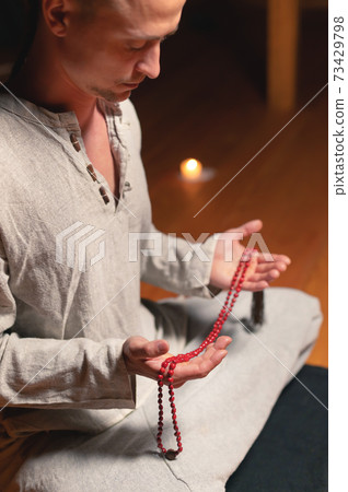 Young attractive man in natural clothes for practice sits in lotus position in a dark room for meditation with rosary in his hands Young attractive man in natural clothes for practice sits in lotus position in a dark room for meditation with rosary in his hands 73429798