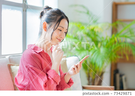 A woman who operates a smartphone while relaxing on the sofa. 73429871
