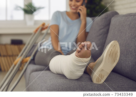 Happy woman with leg in cast sitting on sofa at home and talking on mobile phone Happy woman with leg in cast sitting on sofa at home and talking on mobile phone 73433349