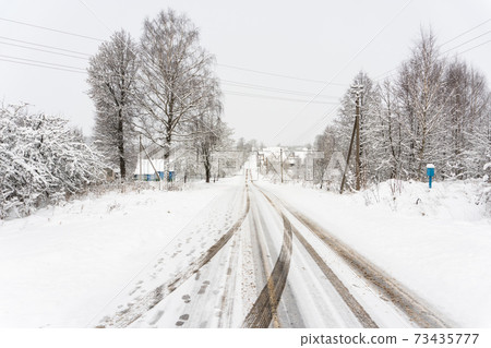 Rural road with a mountain and traces of car tires, poles with power lines and dirty asphalt with salt 73435777