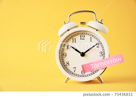 Procrastination, delay and postpone concept. Sticky note with word tomorrow on white alarm clock on yellow background. Urgency time Procrastination, delay and postpone concept. Sticky note with word tomorrow on white alarm clock on yellow background. Urgency time 73436831