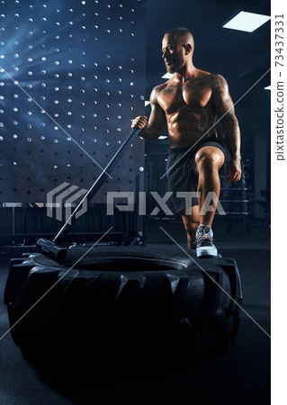 Shirtless bodybuilder carrying sledgehammer on wheel. 73437331