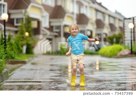 Little boy wearing yellow rubber boots jumping in puddle of water on rainy summer day in small town 73437399
