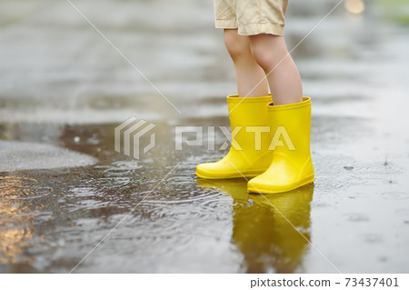 Little boy wearing yellow rubber boots walking on rainy summer day in small town.  73437401