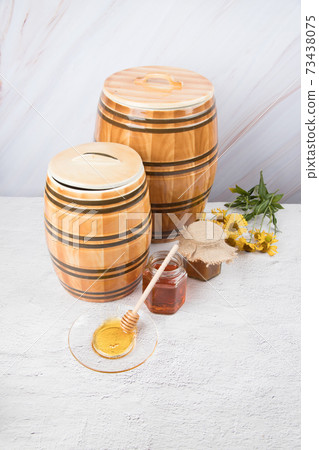 Two ceramic barrels and two glass jars with honey and a saucer with honey and a wooden spoon on a light background. Copy space Two ceramic barrels and two glass jars with honey and a saucer with honey and a wooden spoon on a light background. Copy space 73438075