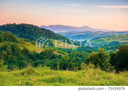 mountainous rural landscape at dawn. beautiful scenery with forests, hills and meadows in morning light. ridge with high peak in the distance. village in the distant valley 73439076