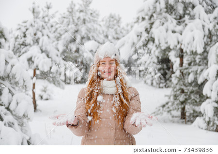 Happy young redhead woman plays with snow in pine tree forest 73439836