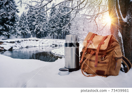 Backpack of traveller, thermos and enameled mug of coffee or tea against beautiful snowy winter landscape background. Backpack of traveller, thermos and enameled mug of coffee or tea against beautiful snowy winter landscape background. 73439981