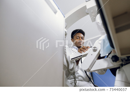 Bottom view of young handsome high-skilled African doctor in white coat, working with modern lithotripter equipment for non-invasive extracorporeal shock wave lithotripsy 73440037