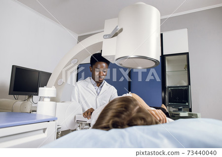 Procedure of extracorporeal shock wave lithotripsy in modern urology medical center. Young African man doctor providing stone treatment or diagnostics for Caucasian female patient using lithotripter Procedure of extracorporeal shock wave lithotripsy in modern urology medical center. Young African man doctor providing stone treatment or diagnostics for Caucasian female patient using lithotripter 73440040