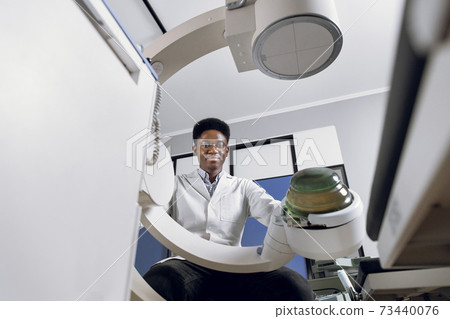 Bottom view of young confident African doctor in white uniform, working with modern lithotripter equipment for treatment of kidney stones. Non-invasive extracorporeal shock wave lithotripsy 73440076