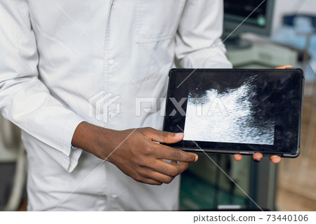 Close up cropped horizontal image of hands of dark skinned male doctor in white coat, holding digital tablet pc with ultrasound scan of the abdomen and kidney Close up cropped horizontal image of hands of dark skinned male doctor in white coat, holding digital tablet pc with ultrasound scan of the abdomen and kidney 73440106