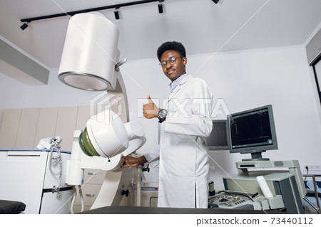 Portrait of handsome young professional African man doctor standing near modern ultrasound and lithotripter machines and showing thumb up. Safety non-invasive stone treatment, shock wave lithotripsy Portrait of handsome young professional African man doctor standing near modern ultrasound and lithotripter machines and showing thumb up. Safety non-invasive stone treatment, shock wave lithotripsy 73440112