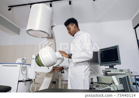 Portait of handsome young confident focused African man doctor, wearing white uniform, working in modern clinic with ultrasonic lithotripter machine. Kidney stones treatment concept Portait of handsome young confident focused African man doctor, wearing white uniform, working in modern clinic with ultrasonic lithotripter machine. Kidney stones treatment concept 73440114