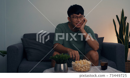 Asian man watching a living room series while eating a popcorn at night. Asian man watching a living room series while eating a popcorn at night. 73449725