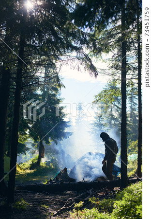 Tent among fir trees under the rays of sun breaking through clouds on cold day. Guy making fire and smoke flows up Tent among fir trees under the rays of sun breaking through clouds on cold day. Guy making fire and smoke flows up 73451759