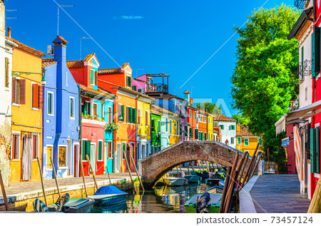 Colorful houses of Burano island. Multicolored buildings on fondamenta embankment of narrow water canal with fishing boats and stone bridge, Venice Province, Veneto Region, Italy. Burano postcard 73457124