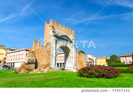 Ruins of ancient brick wall and stone gate Arch of Augustus Arco di Augusto, green lawn with bush of flowers in old historical touristic city centre Rimini 73457141