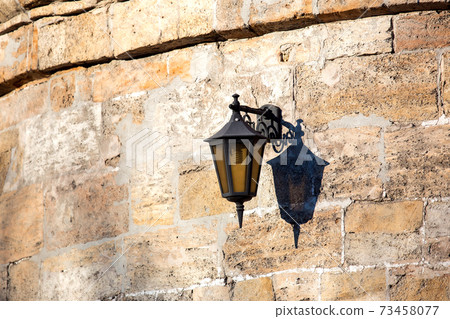 a hinged streetlight lantern from black iron on the background of a stone wall of shell rock. a hinged streetlight lantern from black iron on the background of a stone wall of shell rock. 73458077