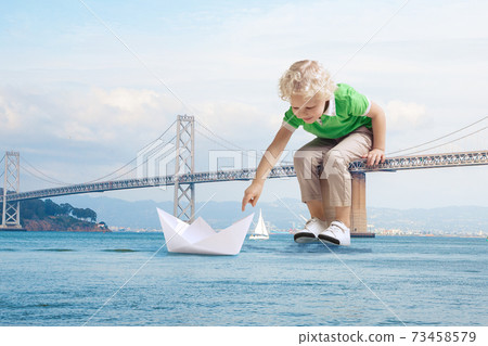 See the world by children's eyes - little cute curly boy playing with paper ship sitting on bridge See the world by children's eyes - little cute curly boy playing with paper ship sitting on bridge 73458579