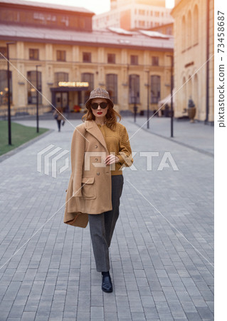 Confident fashion model young woman in sunglasses and hanging on a shoulder autumn beige coat walking on the street happily posing for the camera. Tinted photo Confident fashion model young woman in sunglasses and hanging on a shoulder autumn beige coat walking on the street happily posing for the camera. Tinted photo 73458687