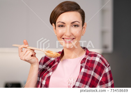 Close up. Charming young housewife with a short hairs cooking a dinner for family trying it using a long wooden spoon standing in the modern kitchen. Healthy food at home 73458689