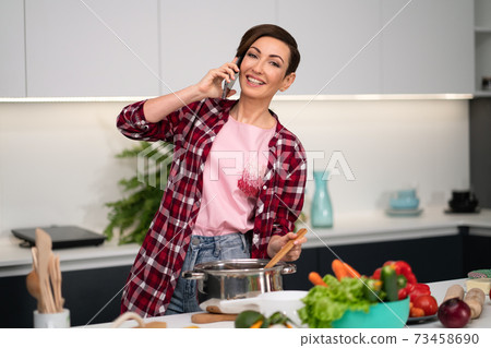 Mixing a soup or ragout while talking on the phone housewife with a short hairstyle. Cooking lunch for family in the modern kitchen. Healthy food at home. Healthy food leaving concept Mixing a soup or ragout while talking on the phone housewife with a short hairstyle. Cooking lunch for family in the modern kitchen. Healthy food at home. Healthy food leaving concept 73458690