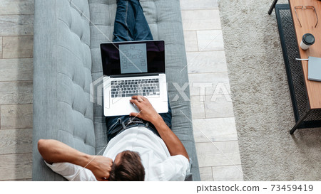 male freelancer is working on a laptop while lying on the couch . 73459419