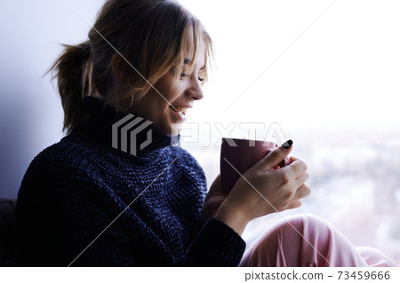 A thoughtful girl in a blue sweater sits by the window with a Cup of coffee A thoughtful girl in a blue sweater sits by the window with a Cup of coffee 73459666