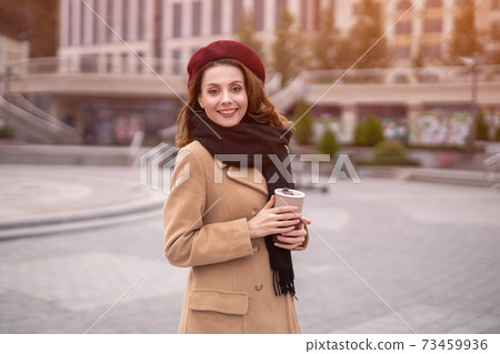 Parisian young woman holding reusable mug with coffee standing outdoors. Portrait of stylish young woman wearing autumn coat and red beret outdoors. Autumn accessories Parisian young woman holding reusable mug with coffee standing outdoors. Portrait of stylish young woman wearing autumn coat and red beret outdoors. Autumn accessories 73459936