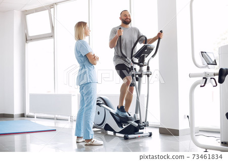 Patient doing exercise on spin bike in gym with therapist 73461834