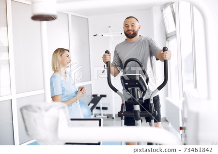 Patient doing exercise on spin bike in gym with therapist Patient doing exercise on spin bike in gym with therapist 73461842