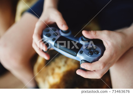 A teenager is sitting on the cofa and playing a 73463676