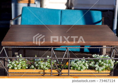 Designer flowerpot made of iron tubular frame with green flowers and white blossoms at the brown wooden table with blue chair of an outdoor cafe, close up nobody. Designer flowerpot made of iron tubular frame with green flowers and white blossoms at the brown wooden table with blue chair of an outdoor cafe, close up nobody. 73467114