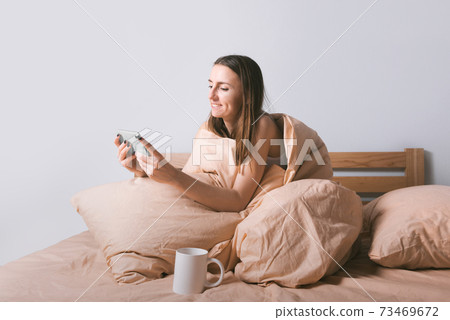 Young smiling woman enjoying using smartphone while lying in comfort bed in the morning 73469672
