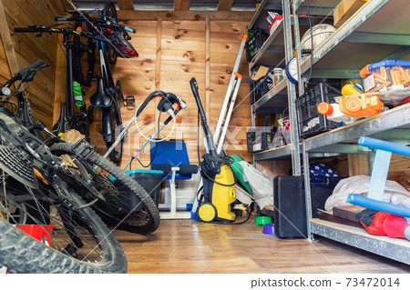 Suburban home wooden storage utility unit shed with miscellaneous stuff on shelves, bikes, exercise machine, ladder, garden tools and equipment. Messy and chaos at house yard barn. Organization order Suburban home wooden storage utility unit shed with miscellaneous stuff on shelves, bikes, exercise machine, ladder, garden tools and equipment. Messy and chaos at house yard barn. Organization order 73472014