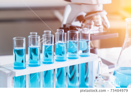 Test tubes with blue liquid in holder with microscope in background. Science glass equipment in laboratory for chemistry and medical research. Test tubes with blue liquid in holder with microscope in background. Science glass equipment in laboratory for chemistry and medical research. 73484979