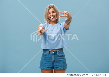 Beautiful tattooed girl enjoying weekends sticking out tongue joyfully winking at camera and smiling showing peace or victory gesture with pulled arms feeling happy posing over blue background Beautiful tattooed girl enjoying weekends sticking out tongue joyfully winking at camera and smiling showing peace or victory gesture with pulled arms feeling happy posing over blue background 73487101