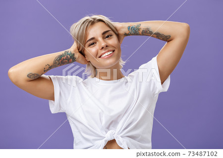 Close-up shot of good-looking stylish and relaxed blond woman with tattoos on arms holding hands behind head smiling with carefree pleased look enjoying weekends over purple background Close-up shot of good-looking stylish and relaxed blond woman with tattoos on arms holding hands behind head smiling with carefree pleased look enjoying weekends over purple background 73487102