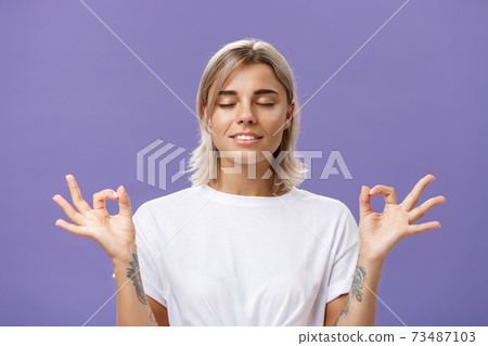 Peaceful calm good-looking female blonde in white t-shirt closing eyes smiling relieved and happy feeling satisfied with life standing in lotus pose with zen gesture over purple background Peaceful calm good-looking female blonde in white t-shirt closing eyes smiling relieved and happy feeling satisfied with life standing in lotus pose with zen gesture over purple background 73487103