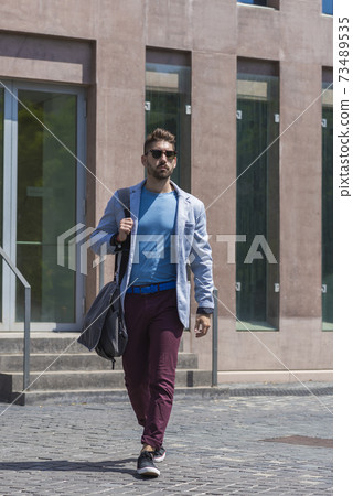 Young businessman walking next to office buildings while holding a shoulder bag outdoors Young businessman walking next to office buildings while holding a shoulder bag outdoors 73489535
