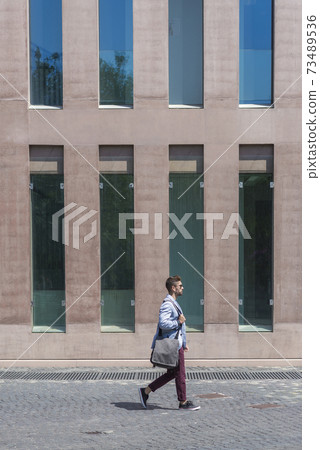 Young businessman walking next to office buildings while holding a shoulder bag outdoors Young businessman walking next to office buildings while holding a shoulder bag outdoors 73489536