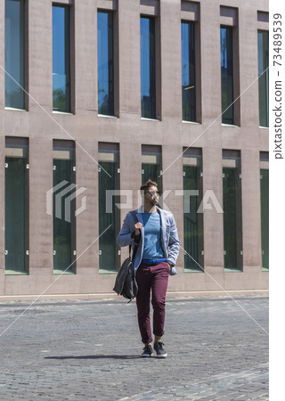 Young businessman walking next to office buildings while holding a shoulder bag outdoors Young businessman walking next to office buildings while holding a shoulder bag outdoors 73489539